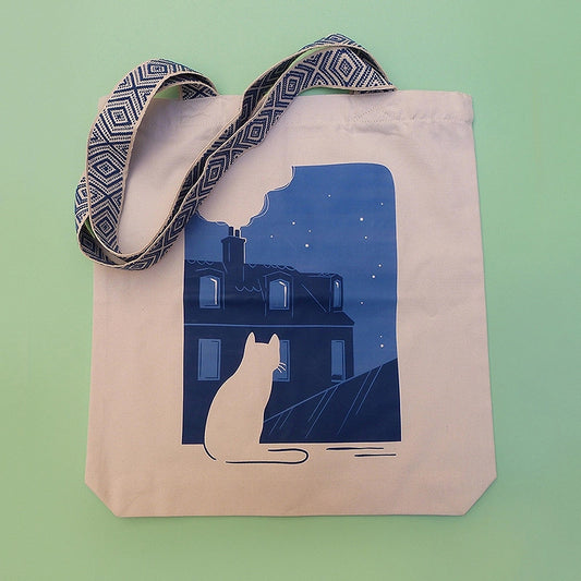 Grand totebag fabriqué en matières recyclées. Poche zippée à l'intérieur. Anses larges et confortables. Impression française. Illustration d'un chat regardant les toits de Paris la nuit.