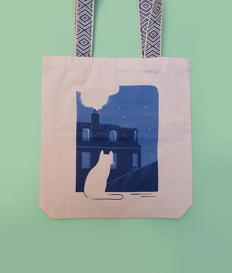 Grand totebag fabriqué en matières recyclées. Poche zippée à l'intérieur. Anses larges et confortables. Impression française. Illustration d'un chat regardant les toits de Paris la nuit.