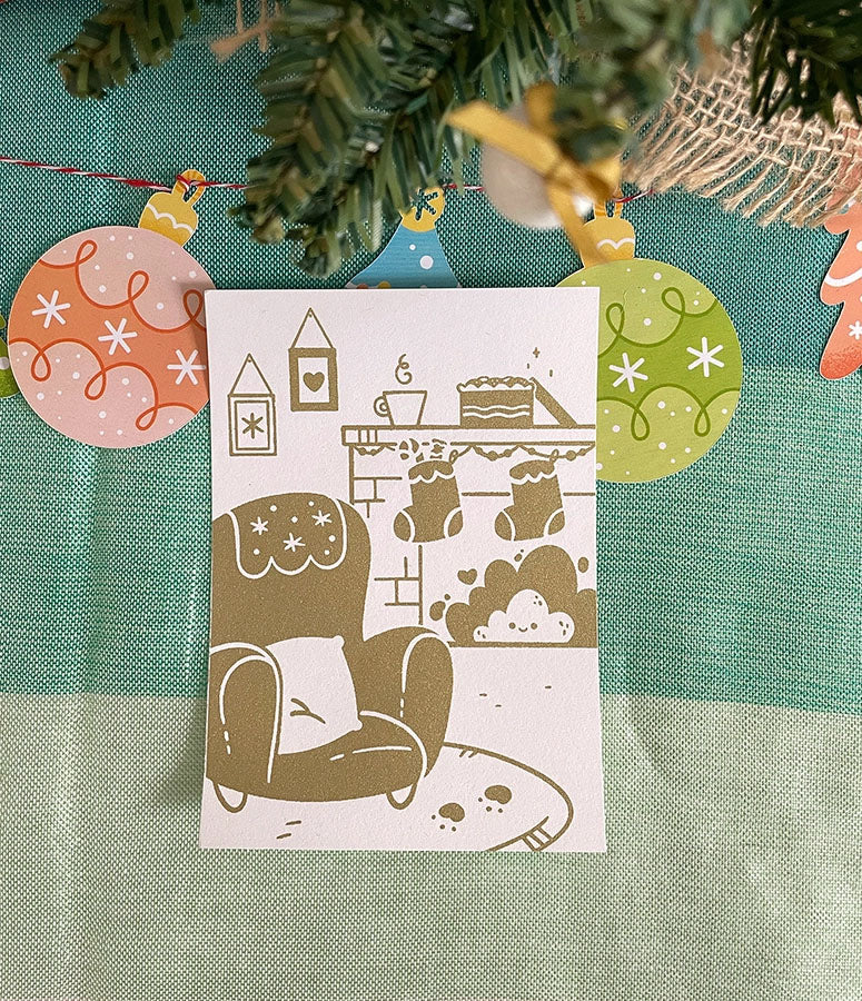 Carte de vœux sérigraphiée artisanalement. Décor cosy de fêtes. Cheminée, biscuits, chocolat chaud, sapin et cadeaux.