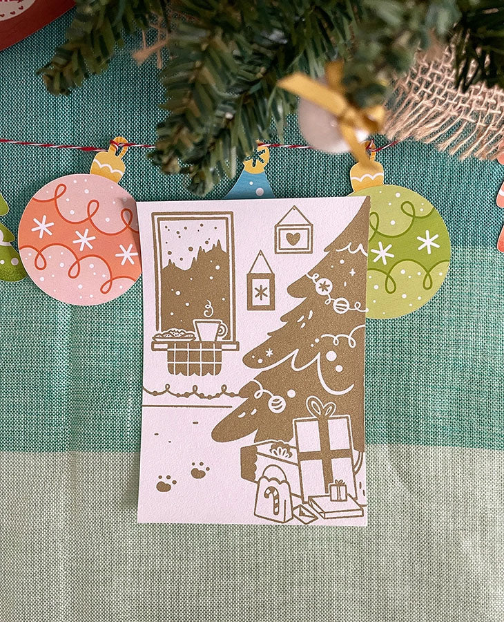 Carte de vœux sérigraphiée artisanalement. Décor cosy de fêtes. Cheminée, biscuits, chocolat chaud, sapin et cadeaux.